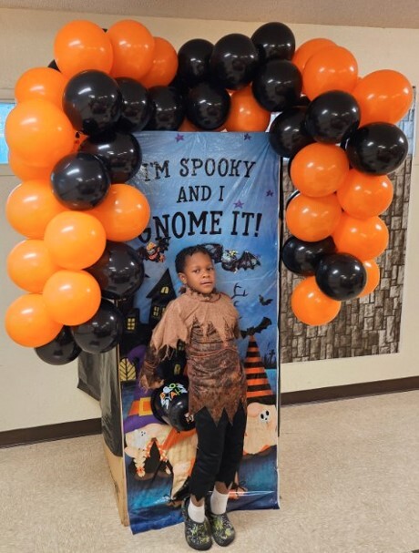 A child dressed up for halloween in front of the halloween sign surrounded by balloons.