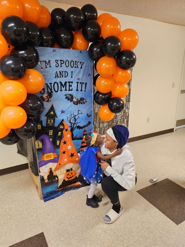 A lady with a little girl in a superhero costume in front of a halloween sign surrounded by balloons.