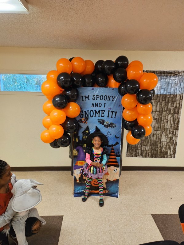 A little girl in a Witch costume in front of a halloween sign surrounded by balloons.