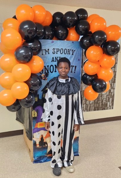 Young man dressed as a clown standing in front of a halloween sign surrounded by balloons.